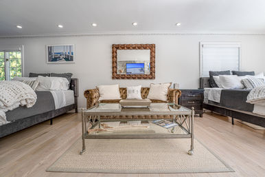 Bright coastal-inspired guest suite with a tufted brown velvet sofa, glass coffee table on a woven rug, twin beds with chunky white knit throws, hardwood floors, decorative wooden mirror and recessed ceiling lights