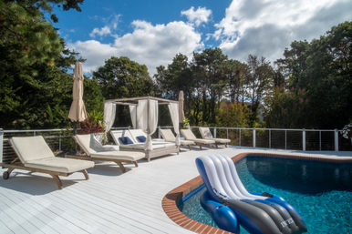Sunny private backyard pool deck with white wood decking, cushioned lounge chairs, a draped canopy daybed, beige umbrellas and a blue inflatable lounger floating in clear water, framed by trees under a blue sky with puffy clouds.