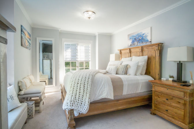 Cozy bright bedroom with rustic wooden bed and headboard, white linens and chunky knit throw, matching wooden nightstand, soft gray walls and a window with blinds letting in natural light