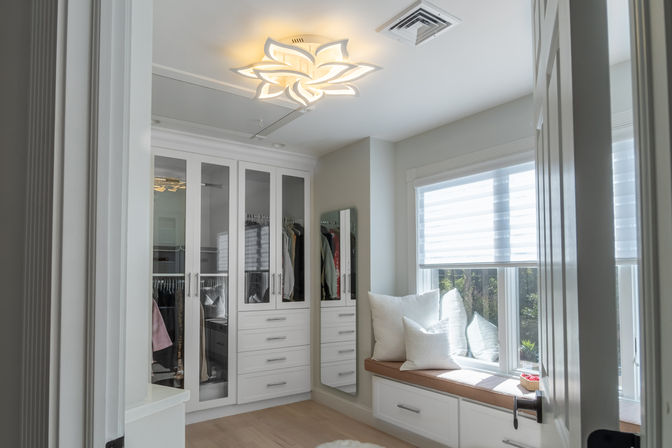 Bright modern walk-in closet with white built-in cabinetry and glass doors, stacked drawers, full-length mirror and hardwood floor, cushioned window seat with white pillows under a large window with zebra shades, lit by a decorative flower-shaped ceiling light.