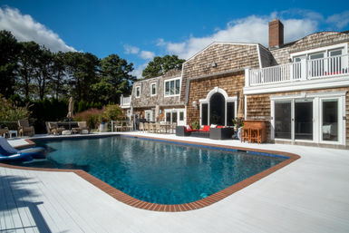 Shingle-style coastal house with large backyard swimming pool, white painted deck, brick coping, outdoor lounge seating and blue sky
