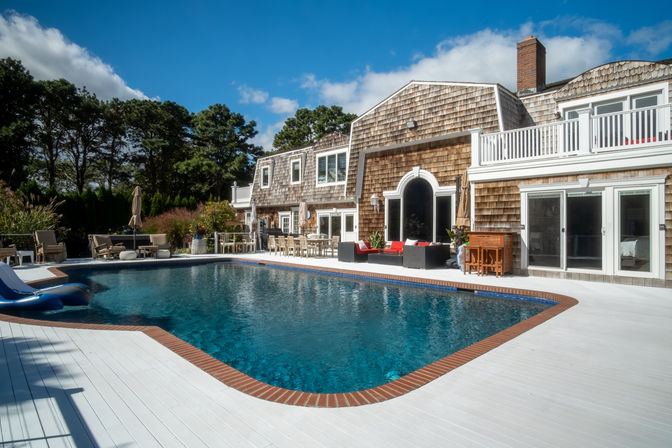 Shingle-style coastal house with large backyard swimming pool, white painted deck, brick coping, outdoor lounge seating and blue sky