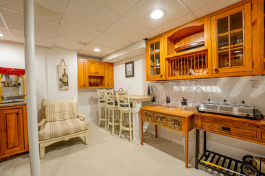 Cozy finished basement home bar with honey pine cabinets, white bar counter and three stools, cushioned bench, clear beverage dispensers, warming trays and popcorn machine under a drop ceiling