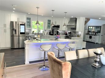 Bright modern white kitchen with large island and blue LED underlighting, stainless steel appliances, pendant lights, three white bar stools, subway tile backsplash and open-concept dining area with dark wood table and upholstered chairs