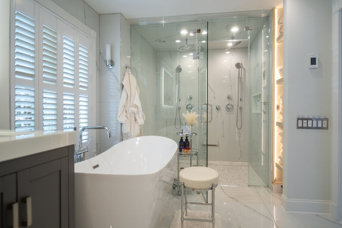 Bright modern luxury bathroom with a white freestanding tub, chrome floor faucet, glass-enclosed dual rain-shower, marble-look tile, plush robe on a hook, illuminated towel shelves and a small stool — spa-like retreat.