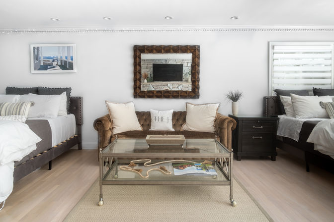 Bright coastal-inspired bedroom lounge with twin beds, tufted velvet sofa, glass coffee table and ocean print on white walls