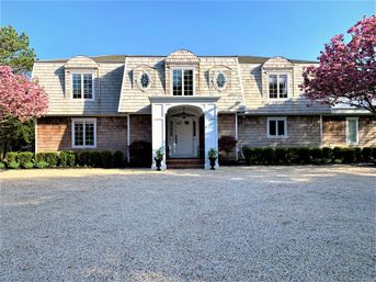 Shingle-style two-story house with white columned portico and oval dormer windows, flanked by cheerful pink flowering trees, low hedges and a wide gravel driveway under a clear blue spring sky.