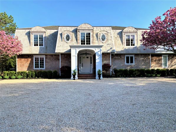 Shingle-style two-story house with white columned portico and oval dormer windows, flanked by cheerful pink flowering trees, low hedges and a wide gravel driveway under a clear blue spring sky.