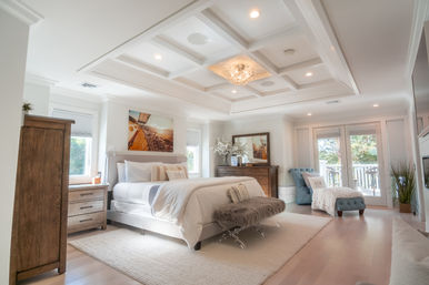 Sunlit coastal-style master bedroom with coffered ceiling and chandelier, king bed with plush fur bench, wooden dressers, tufted chaise, hardwood floors and French doors to a balcony.