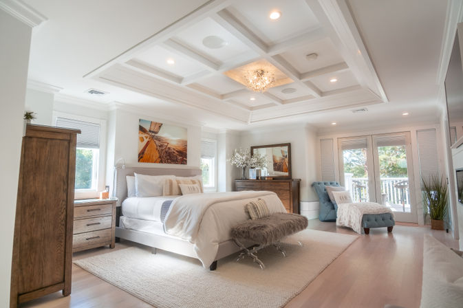 Sunlit coastal-style master bedroom with coffered ceiling and chandelier, king bed with plush fur bench, wooden dressers, tufted chaise, hardwood floors and French doors to a balcony.