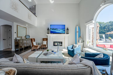 Bright coastal living room with high ceilings, white fireplace and wall-mounted TV, blue surfboard accent, plush gray sofa, blue armchairs and large arched window overlooking a sunny patio and pool