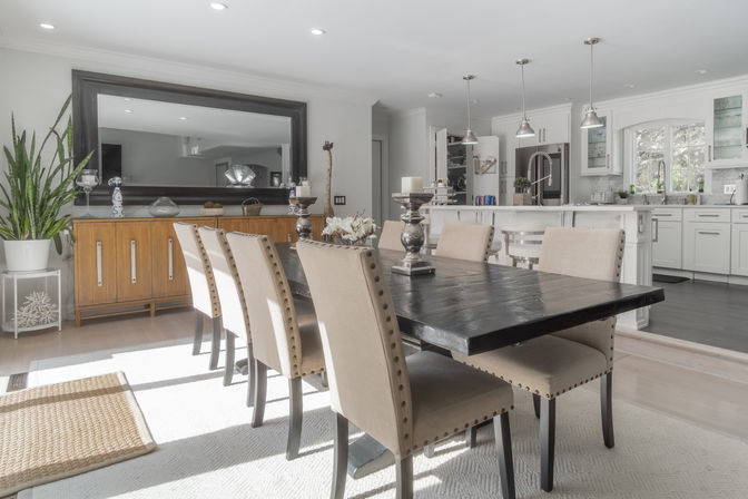 Sunlit open-plan dining room and kitchen in modern farmhouse style featuring a dark wooden dining table with six beige studded chairs, white island with pendant lights, wooden sideboard, large wall mirror and potted plant