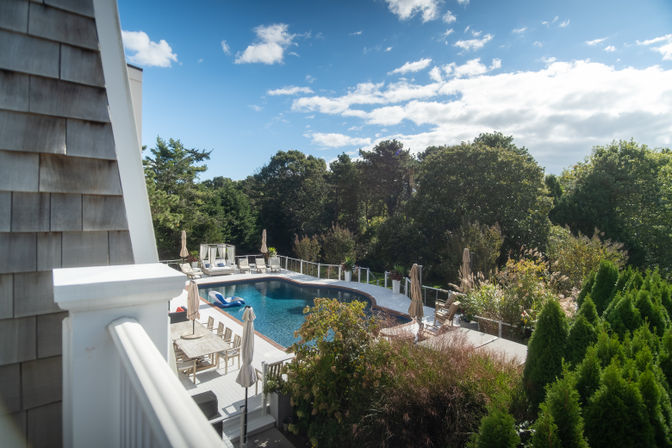 Sunny backyard with an in-ground pool, white wooden deck with dining table, lounge chairs, umbrellas and a cabana, inflatable float and lush trees surrounding the area