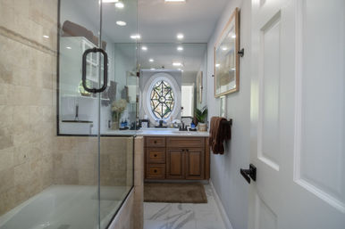 Cozy residential bathroom with glass-enclosed tub-shower, beige stone tiles, marble-look floor, wooden vanity under an oval decorative window, framed art and brown towels.