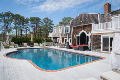 Shingle-style seaside home with a large blue swimming pool, white wooden deck, lounge chairs, umbrellas and red patio seating under a bright sky