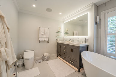 Bright, spa-like modern master bathroom with freestanding white soaking tub, gray double vanity with under-cabinet lighting and large backlit mirror, chrome faucets, wall-mounted towel rack with white towels, plush rugs and shuttered window.