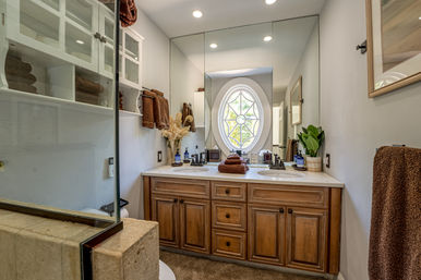 Bright bathroom with double-sink wood vanity and white countertop, stacked brown towels, large mirrored wall, oval decorative window with star pattern, glass shower partition and potted plant.