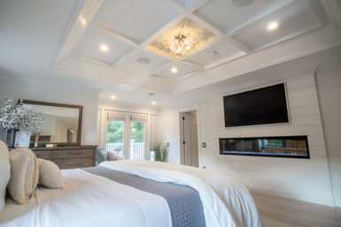 Bright modern master bedroom with coffered ceiling and crystal chandelier, wall-mounted TV above a linear electric fireplace, plush white bedding, wooden dresser with mirror, and glass doors opening to a balcony with greenery.