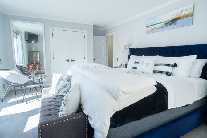 Sunlit coastal-inspired bedroom with navy tufted headboard, crisp white linens, striped accent pillows, tufted bench at the foot, and a small sitting nook with black wire chairs and red flowers.