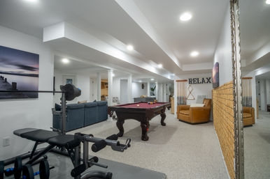 Finished basement recreation room with burgundy pool table, home gym bench and dumbbells, blue sectional sofa and tan leather armchair beneath a 'RELAX' wall sign, neutral carpet and recessed lighting.