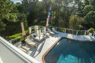 Sunlit backyard swimming pool with brick-trimmed edge, white wood deck, canopy daybed and cushioned lounge chairs surrounded by trees and lawn