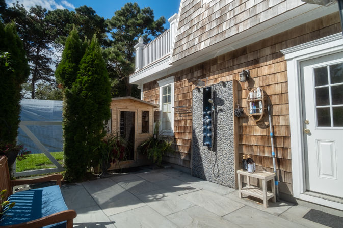 Sunny backyard patio with cedar-shingle siding, pebble-tiled outdoor shower panel, small wooden shed, potted plants and blue-cushioned bench.