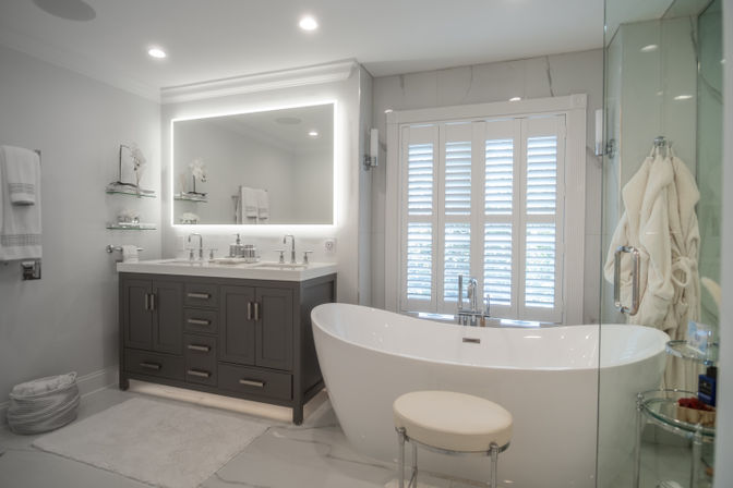 Spa-like bright modern bathroom with freestanding white soaking tub, gray double-sink vanity under LED-lit mirror, plantation shutters and glass shower