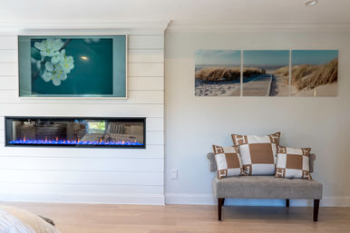 Bright coastal-style living room corner with white shiplap wall, long electric fireplace with blue flames, teal floral art above, beach boardwalk triptych, and upholstered bench with tan-and-white check pillows.