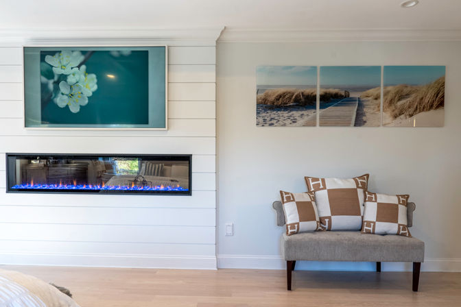 Bright coastal-style living room corner with white shiplap wall, long electric fireplace with blue flames, teal floral art above, beach boardwalk triptych, and upholstered bench with tan-and-white check pillows.