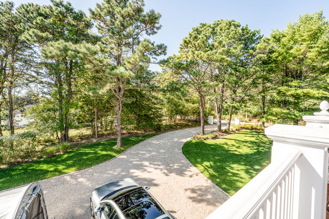 Sunny balcony view of a curved gravel driveway with a parked car, manicured green lawn and tall pine trees lining a suburban yard.