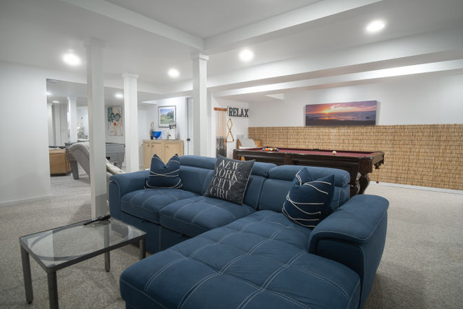 Cozy finished basement game room with a blue sectional sofa, glass coffee table, pool table, recessed lighting and wall art.