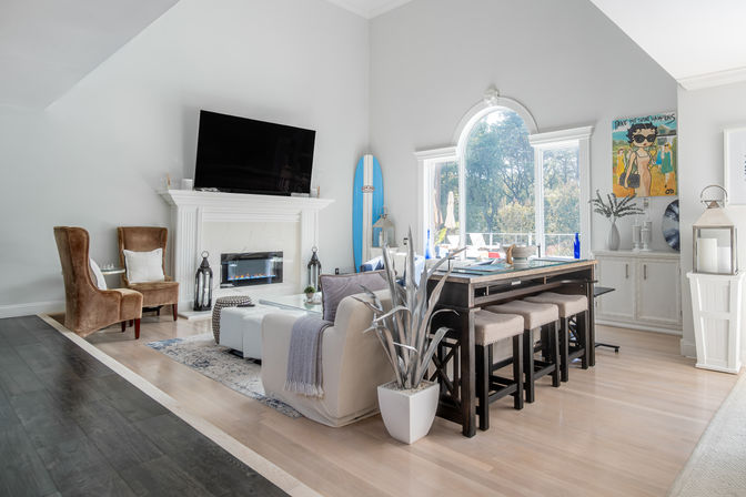 Bright modern coastal-style living room with vaulted ceiling, white fireplace and TV, arched window overlooking trees, blue surfboard, cozy seating, and dark wood high-top table with stools.
