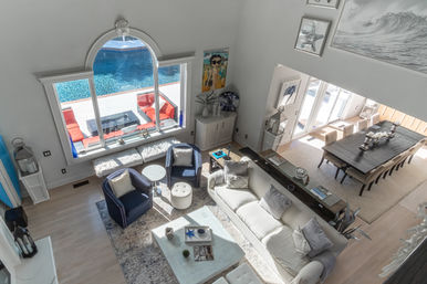 Sunlit coastal living room with high ceilings and large arched window overlooking a blue backyard pool and red patio seating, featuring a neutral sofa, navy accent chairs, marble coffee table and open-plan dining table.