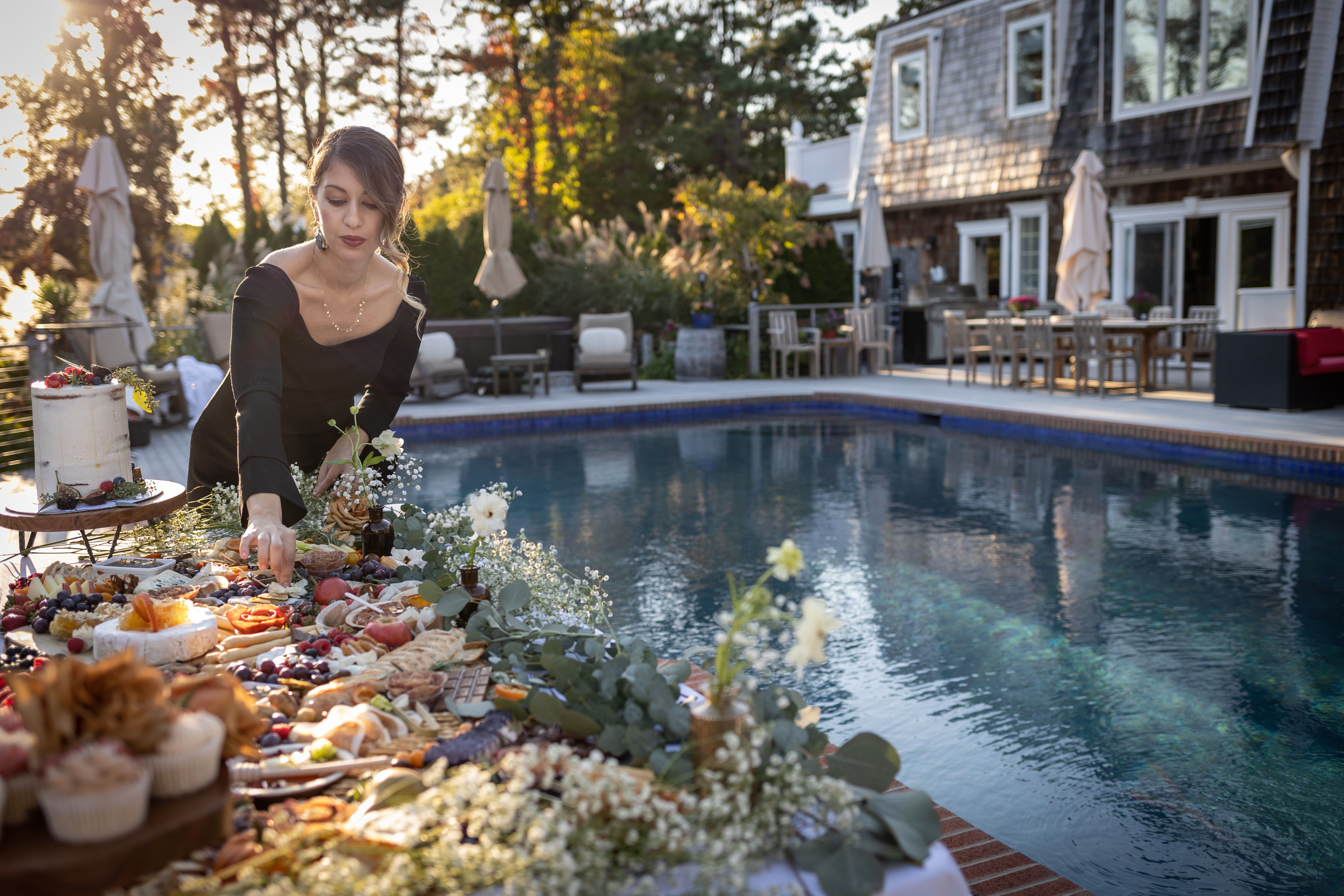 Person arranging an abundant charcuterie and dessert spread on a table beside a backyard swimming pool at sunset, with patio seating and a shingled house in the background.