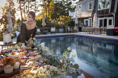 Person arranging an abundant charcuterie and dessert spread on a table beside a backyard swimming pool at sunset, with patio seating and a shingled house in the background.
