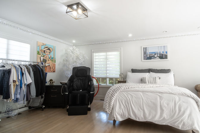 Bright, airy modern bedroom with white bedding and chunky knit throw, black massage chair, clothing rack of hanging shirts, hardwood floors, window with blinds and coastal wall art.