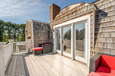 Sunny coastal-style rooftop deck with weathered wood shingles, white railing, glass French doors, brick chimney and wicker seating with bright red cushions, trees and blue sky beyond