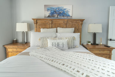 Cozy modern farmhouse bedroom with carved wooden headboard, crisp white bedding and chunky knit throw, textured decorative pillows, matching wooden nightstands with black table lamps, and vibrant blue tree wall art above the bed.