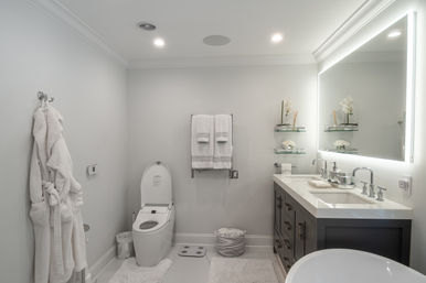 Spa-like modern white residential bathroom with LED backlit mirror, gray double-sink vanity, sleek electronic toilet, glass shelves with orchids, plush robe and towels, and neutral minimalist decor.