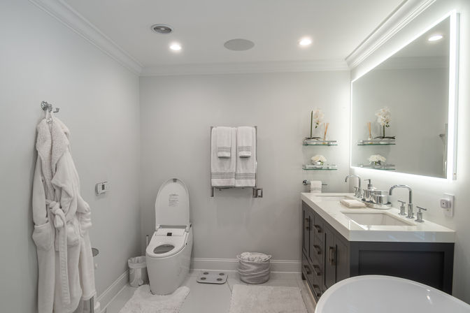 Spa-like modern white residential bathroom with LED backlit mirror, gray double-sink vanity, sleek electronic toilet, glass shelves with orchids, plush robe and towels, and neutral minimalist decor.