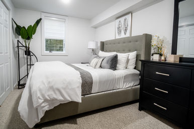 Cozy modern bedroom interior with a tufted gray headboard, crisp white bedding and gray accent pillows, black dresser with mirror and orchid, tall potted plant, bedside lamp and small window.
