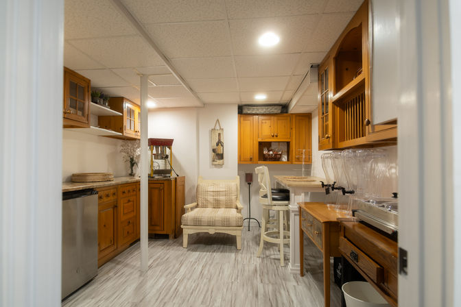 Cozy finished basement kitchenette and wet bar with honey-toned wooden cabinets, marble-look countertop, stainless mini-fridge, cream upholstered armchair, white bar stools, clear beverage dispensers, and gray wood-look flooring.