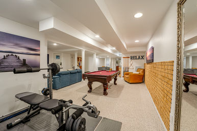 Finished basement recreation room with red-felt pool table, home gym bench and weights, blue sectional sofa, leather armchair, wall art and a 'RELAX' sign under recessed lighting.