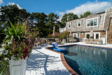 Sunny backyard pool deck with a blue inflatable lounger in a brick-coped pool, white painted decking, shingle-style house, patio dining set, lounge chairs and colorful flowering planters