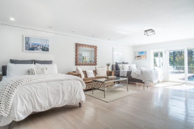Bright, airy poolside master suite with white bedding, two beds, tufted velvet sofa, glass coffee table, hardwood floors and sliding glass doors opening to a sunlit patio and pool