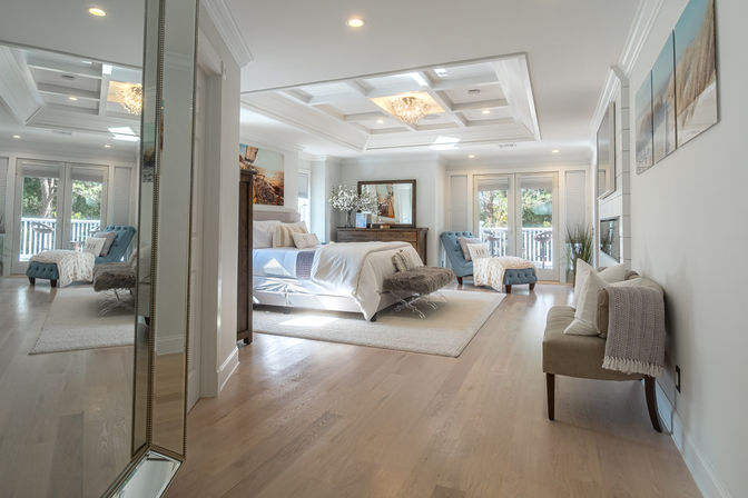 Sunlit spacious master bedroom with coffered ceiling and chandelier, king bed with white linens, tufted blue lounge chairs, mirrored dresser, hardwood floors and French doors opening to a tree-lined balcony.