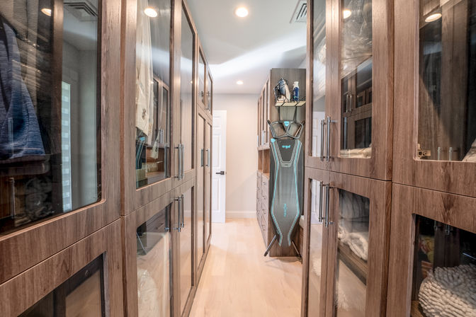 Modern walk-in closet with floor-to-ceiling walnut cabinets and glass-front doors, recessed lighting, organized shelving and a standing ironing board