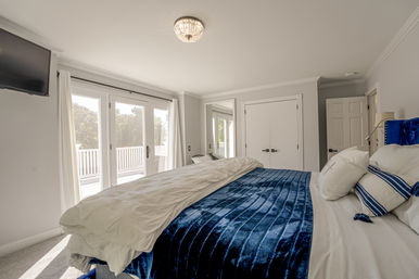 Bright master bedroom with navy velvet bedspread and white linens, tufted blue headboard, wall-mounted TV, double closet and crystal ceiling light, French doors opening to a sunlit deck.