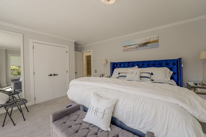 Coastal-inspired master bedroom with king bed in white linens, bold blue tufted velvet headboard, beach wall art, mirrored nightstand, tufted bench and soft gray carpet.