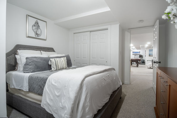 Finished basement bedroom with gray upholstered bed, white-and-gray layered bedding and decorative pillows, framed seashell art, sliding closet doors, and an open door revealing a rec room with a pool table.